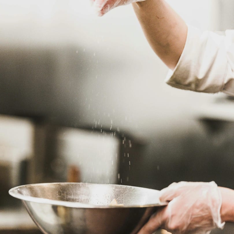 A chef's gloved hand sprinkles ingredients into a metal mixing bowl in a kitchen.