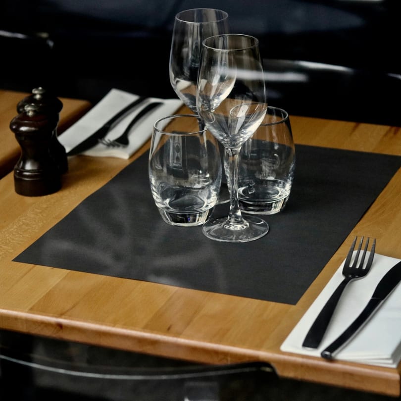 A restaurant table set with wine glasses, water glasses, and cutlery on a black placemat.