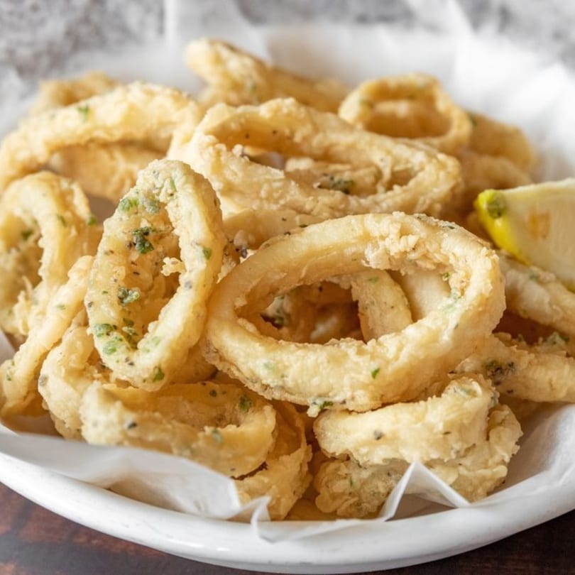 Crispy fried calamari rings with herbs and a lemon wedge in a white bowl.