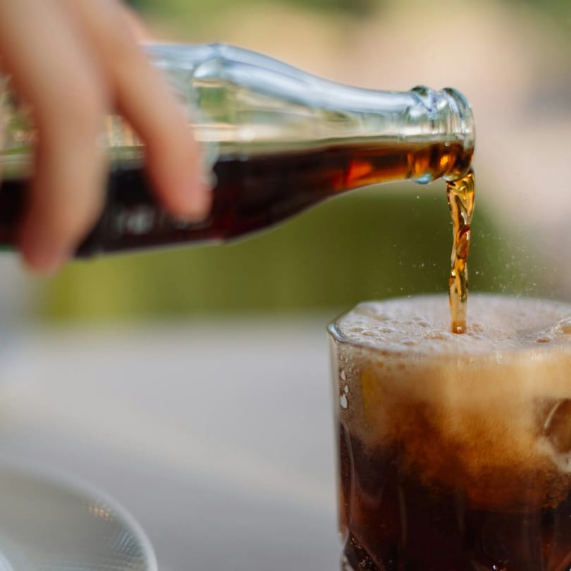 A hand pours dark soda from a glass bottle into a glass with ice, creating foam.