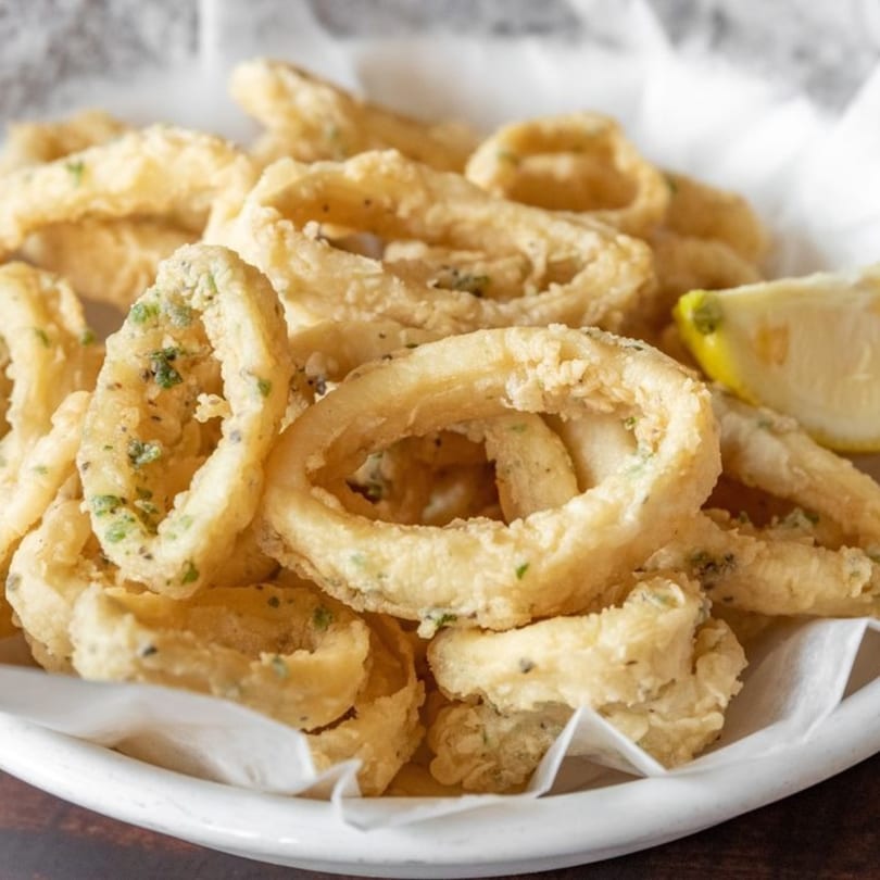 Crispy fried calamari rings with herbs and a lemon wedge, served in a white bowl.
