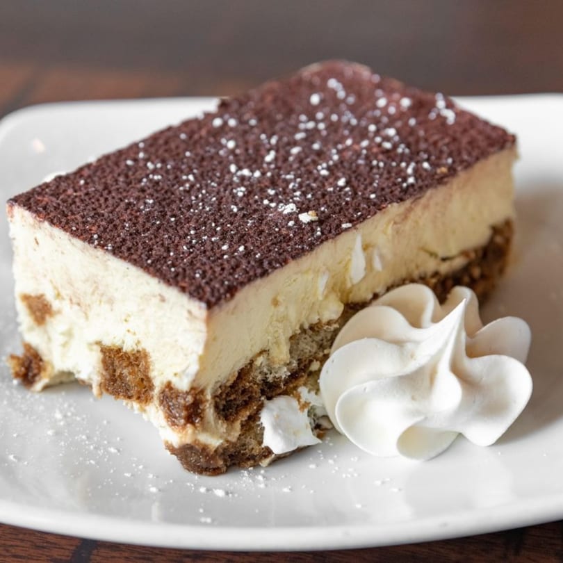 A slice of tiramisu with cocoa powder and powdered sugar, served with whipped cream.