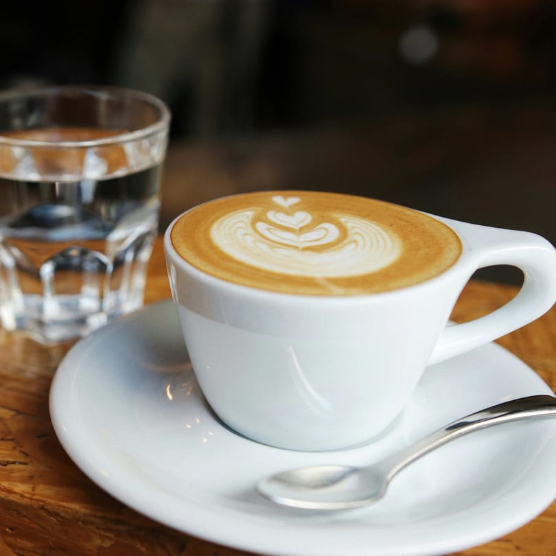 A white cup of latte with heart-shaped foam art, a spoon, and a glass of water.
