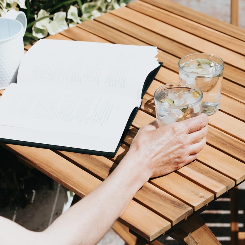 A person's hand holds a glass of iced water next to an open book on a wooden table.